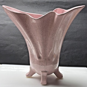 Puede incluir: Un jarrón de cerámica rosa con una textura moteada y una forma acampanada asimétrica. El jarrón tiene tres patas cortas y un borde ondulado. El color es un rosa suave y apagado.