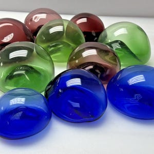 May include: A collection of colorful, smooth glass stones in shades of blue, green, red, and purple. The rounded shapes reflect light, creating highlights. These decorative stones could be used as vase fillers or home decor accents.