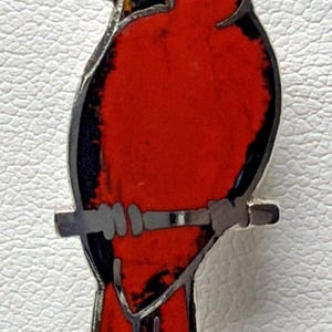 Puede incluir: Un broche de metal plateado con forma de ave cardenal. El pájaro presenta secciones incrustadas de color rojo vibrante en el cuerpo, la cabeza y la cola, con detalles negros alrededor del ojo y el ala, y un pequeño detalle naranja en el pico.