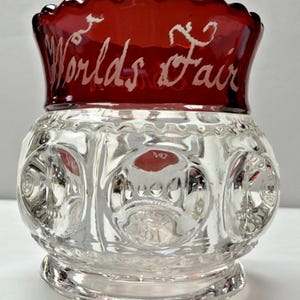以下が含まれることがあります： 赤い縁が付いた透明なガラス瓶。瓶には装飾的な模様があり、「Worlds Fair」の文字が赤い縁に白い筆記体で書かれています。瓶にはスカラップエッジがあります。