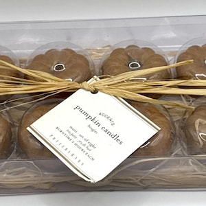 May include: A set of six pumpkin-shaped candles in a clear plastic box with a brown straw-like filler. The candles are brown and have a label that reads "Accents Pumpkin Candles, mini set of six, burn time 2 hours each, Primitives by Kathy."