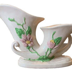 May include: A vintage, off-white ceramic vase with a unique horn-like shape. The vase features embossed pink flowers, green leaves, and stems. The base is round, and the vase has a handle. A decorative piece for home decor.