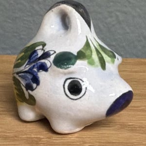 May include: A small, white ceramic figurine of an animal, possibly a pig or a similar creature. It features black, blue, and green painted details, including a dark eye and floral accents. The figurine is on a wooden surface.