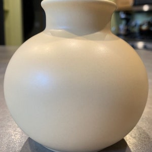 May include: A round, cream-colored ceramic vase with a smooth, glossy finish.