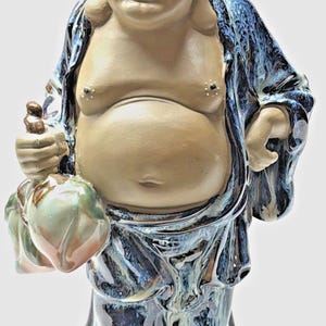May include: A ceramic statue of a smiling Buddha figure. The figure has a large belly and is wearing a blue robe with a glossy finish. The Buddha is holding a bag and a small object in its hands. The statue is tan and blue.