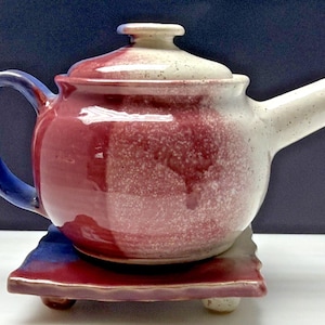 May include: A ceramic teapot with a matching lid and a small, square base. The teapot is primarily burgundy with a white speckled top and spout. The handle is blue. The base is burgundy and blue with small, rounded feet.
