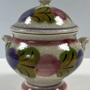 May include: A white ceramic soup tureen with a lid, featuring a hand-painted floral design in shades of purple, pink, green, and yellow. The tureen has two handles and a round base.