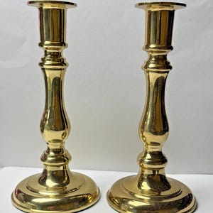 May include: Two polished brass candlesticks. Each candlestick has a round base, a slender, curved stem, and a flat top. The brass has a shiny, reflective surface, and the design is classic and elegant.