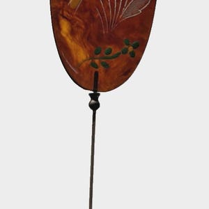 May include: An oval-shaped decorative pin with a brown background and floral design. The pin features a long, slender, dark metal stem. The floral design includes white, yellow, blue, and green accents.
