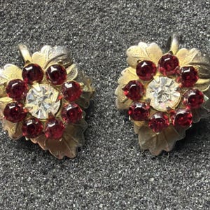 May include: A pair of vintage earrings featuring a leaf design with gold-toned metal and silver-toned accents. Each earring has a central clear rhinestone surrounded by smaller, deep red rhinestones. The earrings are set against a textured gray background.