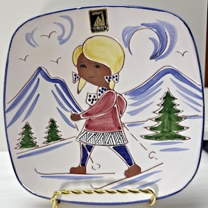 May include: Square ceramic plate with a hand-painted design of a skier in a red jacket and blue trousers, skiing down a snowy mountain. The plate has a white background with blue and green accents. The plate is 20 cm square.