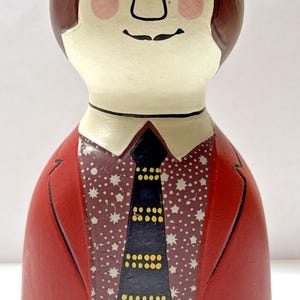 May include: A hand-painted ceramic figurine of a man with brown hair and a red suit. The figurine has a white face with pink cheeks, a black mustache, and a black tie with yellow dots. The shirt has a white star pattern.