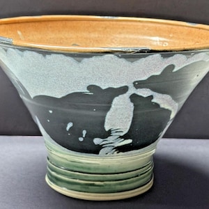 May include: A wide, flared ceramic bowl with a tan interior and a dark green exterior featuring a white, cloud-like glaze pattern. The base is narrow with horizontal ridges, and the rim is smooth and rounded, showcasing a hand-thrown pottery design.