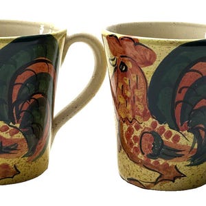 May include: Two ceramic mugs with a rooster design. The mugs have a yellow background with a rooster illustration in red, orange, and green. Each mug has a beige handle and rim.