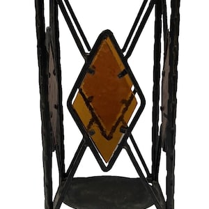 May include: Black wrought iron candle holder with geometric diamond-shaped amber glass inserts. The decorative stand has a small circular base and a larger, open top. The metalwork features a textured finish.