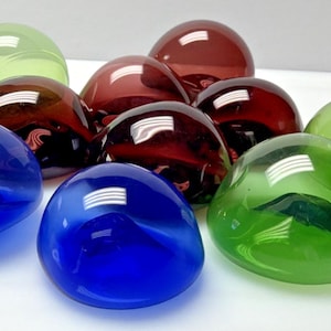 Puede incluir: Una colección de objetos de vidrio lisos y redondeados en varios colores, incluyendo azul, verde y burdeos. Las piezas transparentes reflejan la luz, creando reflejos. Los objetos parecen ser elementos decorativos o artísticos.