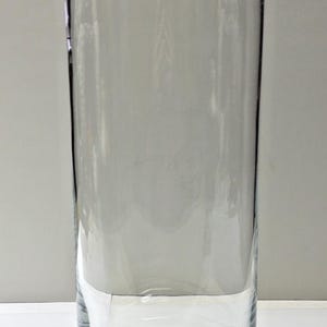 May include: A tall, clear cylindrical glass vase with a thick, weighted base. The upper portion of the glass features a small, square white sticker with the black text LSA INTERNATIONAL printed on it. The vase has a smooth, transparent surface.
