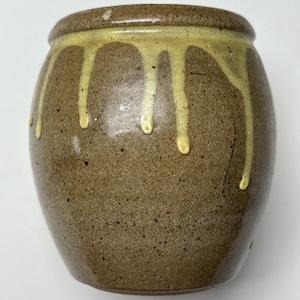 May include: A small, round, brown ceramic jar with a speckled texture. The rim and upper portion of the jar are adorned with a light yellow glaze that drips down the sides, creating a unique, handcrafted appearance. The jar has a wide opening.