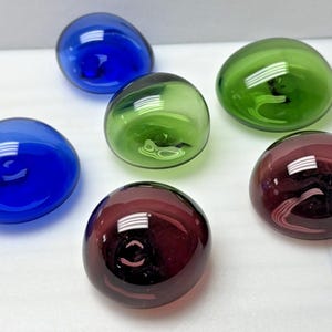 May include: Six colorful glass paperweights in shades of blue, green, and burgundy. Each paperweight has a smooth, rounded shape. The glass reflects light, creating highlights and shadows.