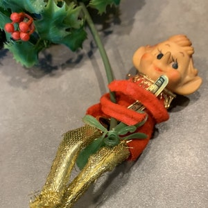 May include: A vintage Christmas elf ornament with a red velvet body, gold glitter legs, and a painted face. The elf is holding a small green sprig of holly.