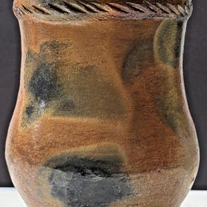 May include: Handmade ceramic vase with a textured surface and a unique, organic design. The vase features a blend of brown, black, and green hues, with a decorative rope-like detail around the rim. A rustic, earthy piece for home decor.