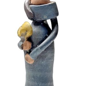 May include: A handcrafted ceramic figurine of a woman in a long, blue dress. The figure holds a golden object. The woman's head and hands are a light brown color. The figurine has a simple, elegant design.