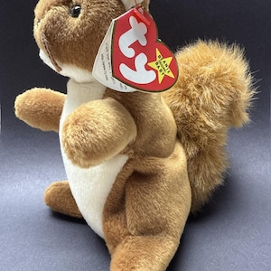 May include: Brown and white plush squirrel with a long bushy tail. The squirrel is holding a red Ty Beanie Baby tag with a white star and the words "Beanie Original Baby" on it.