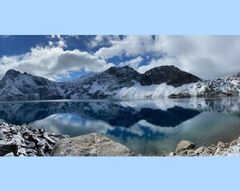 Op de afbeelding: Een panoramisch uitzicht op een besneeuwde bergketen die wordt weerspiegeld in een stil meer. De lucht is blauw met witte wolken.