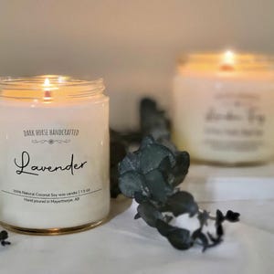 May include: Two lit, white soy wax candles in glass jars. The front candle has the word "Lavender" in script, with text that reads "100% Natural Coconut Soy wax candle | 7.5 oz". The background candle is out of focus.