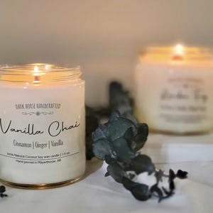 May include: Two lit candles in glass jars. The front candle has a label reading "Vanilla Chai" with the ingredients listed below. The candle is 213 grams. The second candle is out of focus, but has a lit flame and a label.