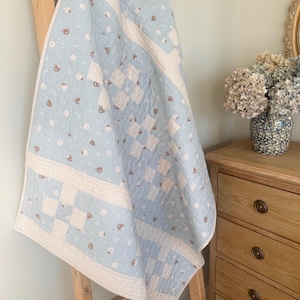 May include: A light blue and white quilt with a repeating pattern of teddy bears and geometric shapes. The quilt is draped over a wooden ladder, showcasing its design and texture. The quilt's dimensions are not visible.