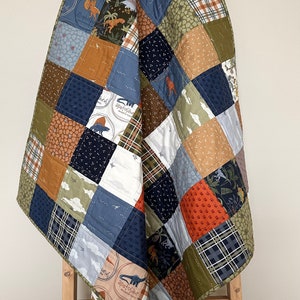 May include: A patchwork quilt with a dinosaur theme, draped over a wooden ladder. The quilt features squares of various patterns and colors, including blue, green, brown, and orange, with dinosaur illustrations and text. The quilt is approximately 60 inches by 60 inches.