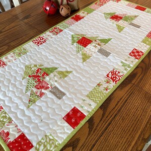 May include: A quilted Christmas table runner featuring a white background with green, red, and gray tree designs. The runner has a green border and is decorated with festive patterns. The runner is approximately 12 inches wide and 36 inches long.