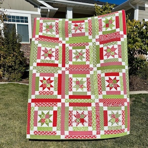 May include: A large, colorful quilt with a repeating pattern of red and green star blocks. The quilt features a patchwork design with various floral and dotted fabrics. The quilt is displayed outdoors, showcasing its vibrant colors and intricate design.