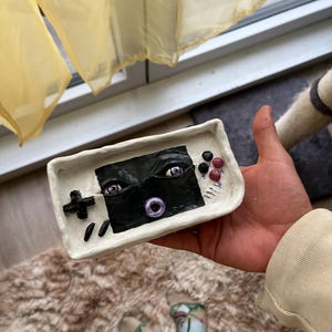 Op de afbeelding: Een handgemaakt, wit keramisch dienblad in de vorm van een retro handheld gameconsole. Het scherm heeft een zwart ontwerp met ogen en een paarse cirkel. Het heeft een zwart kruisvormig D-pad en rode en zwarte knoppen. Het dienblad wordt in een hand gehouden.
