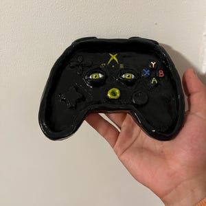 Puede incluir: Un mando de juego de cerámica negro pintado a mano con ojos verdes, una X amarilla y un círculo verde. El mando tiene una estrella, una flor y las letras X, Y, A y B.