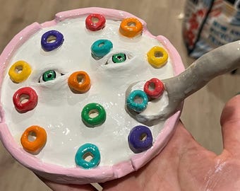 May include: A ceramic bowl with a pink rim, white interior, and colorful cereal pieces. The bowl has two green eyes and a gray spoon with cereal. The cereal pieces are red, orange, yellow, green, blue, and purple.