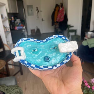 Op de afbeelding: Een hartvormige keramische schaal met een zwembadontwerp. Het zwembad is lichtblauw met twee geschilderde ogen en een blauwe knop. De rand is blauw en wit geblokt. Een wit duikplank en ladder zijn bevestigd.