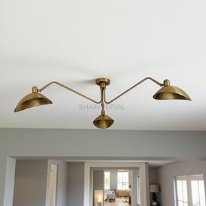 May include: A brass-toned ceiling light fixture with three curved arms and bell-shaped shades. The fixture is mounted on a white ceiling. The text "SHAIMERIAL" is visible on the fixture.