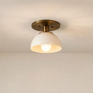 May include: A flush mount ceiling light fixture featuring a circular antique brass canopy and a smooth, white dome-shaped shade. The fixture includes a single exposed light bulb centred within the shade, casting a warm glow downwards.
