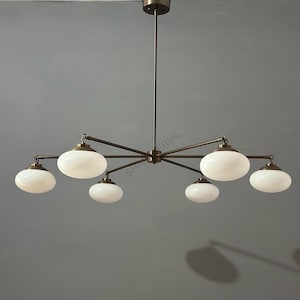 Puede incluir: Un candelabro de color bronce con seis pantallas de vidrio blanco ovaladas. La lámpara tiene un vástago central y brazos radiantes, creando un diseño moderno. El candelabro está suspendido del techo.