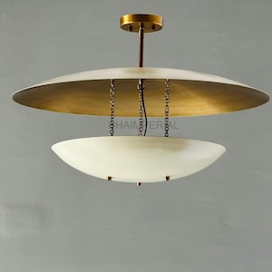 Op de afbeelding: Een hanglamp in mid-century modern stijl. De lamp heeft een grote, witte, schotelvormige kap met een goudkleurige binnenkant. Een kleinere, komvormige kap hangt eronder. De lamp wordt opgehangen aan kettingen en een centrale stang.