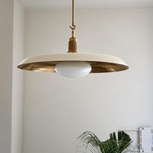 Peut inclure: Une suspension lumineuse dotée d'un large abat-jour plat en forme de soucoupe blanc cassé avec une finition intérieure dorée métallique. Un diffuseur central en verre blanc est suspendu à une tige et une armature en métal laitonné.