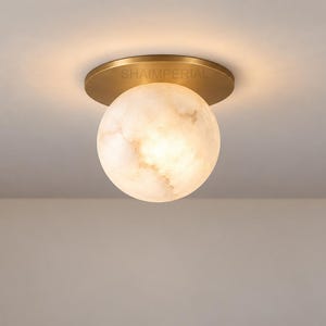 May include: A flush mount ceiling light featuring a spherical, translucent white stone shade with natural veining, attached to a circular brushed brass base. The word SHAIMPERIAL is printed in small, dark letters on the brass mounting plate.