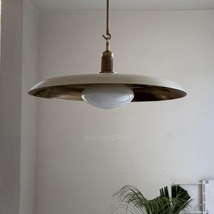 May include: A contemporary pendant light. It features a white, disc-shaped shade with a brass-coloured underside. The light has a frosted glass globe and a wooden and brass stem. The fixture is suspended by a thin wire. The word "SHAIMPERIAL" is printed on the image.