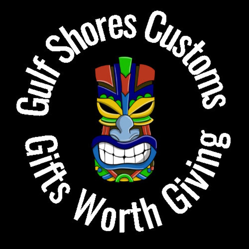 Puede incluir: Ilustraci&oacute;n de una m&aacute;scara tiki colorida con una amplia sonrisa, rodeada por el texto "Gulf Shores Customs Gifts Worth Giving" en disposici&oacute;n circular, sobre fondo negro.