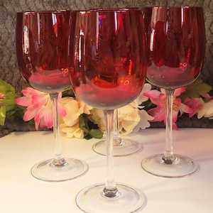 May include: Five ruby red wine glasses with a classic design. Each glass has a rounded bowl and a long, clear stem. The glasses are arranged on a white surface, with flowers in the background.