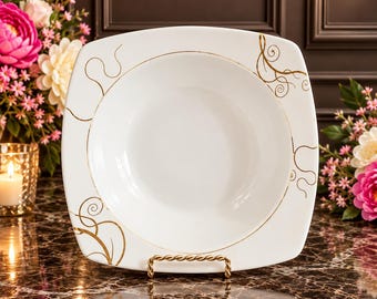 May include: A square white ceramic bowl with rounded corners, featuring delicate gold-colored swirling line patterns on the rim. The bowl is displayed on a gold-toned metal stand, showcasing a smooth, glossy finish and a wide, deep basin.