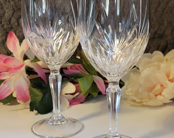 May include: Two clear crystal wine glasses with intricate etched designs. The glasses have long stems and round bases. The background features pink and white flowers and green leaves.