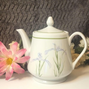 May include: White ceramic teapot with a lid, handle, and spout. The teapot is decorated with a green band around the lid and a floral design featuring blue and purple irises. The teapot is on a white surface.